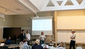 Students presenting in fron of a large TV screen in a high ceiling classroom.