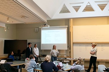 Students presenting in fron of a large TV screen in a high ceiling classroom.