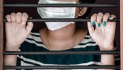 Girl behind bars with a facemask covering her mouth.