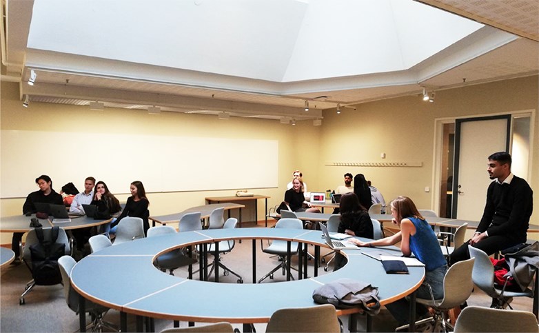 Students sitting in a lecture room at different shaped spread out tables. Some looks happy and from ceiling comes natural daylight.