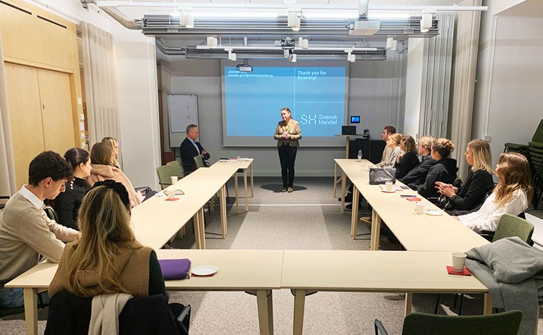 Several students sitting around a u-shaped desk looking towards and listening to a presenter up front. A powepoint slide is projected with a text "thank you for listening" and a with a Svensk Handel logo on it.