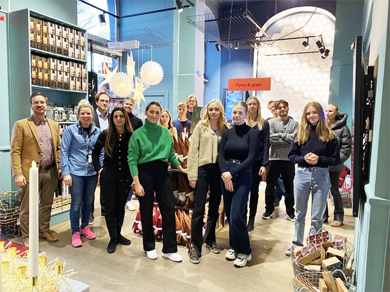 Group of students standing in a group together with representatives from Clas Ohlson in a high ceiling physical store. The people is smiling into the camera. The room / store is decorated with the seasonal christmas items that's for sale.