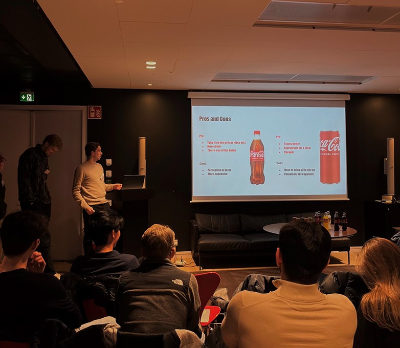 A student presenting in fron of a coca-cola labelled ppt in a dark lit room.