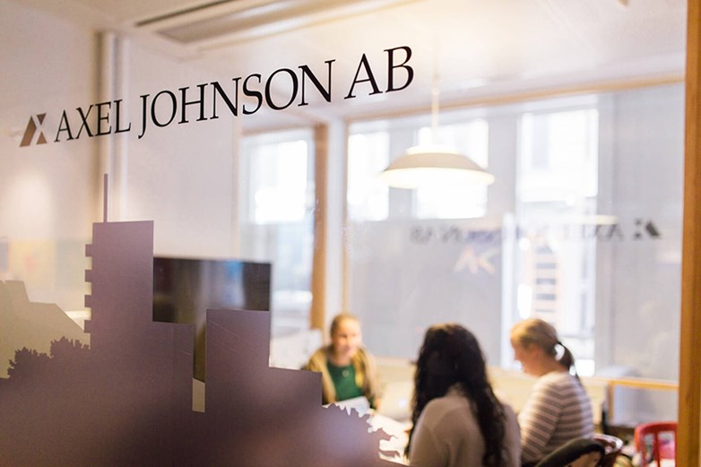 Picture of three persons having a workshop or meeting in a smaller cosy  office space. They are blurred and on transperant glass "Axel Johnson AB" is written in black text.