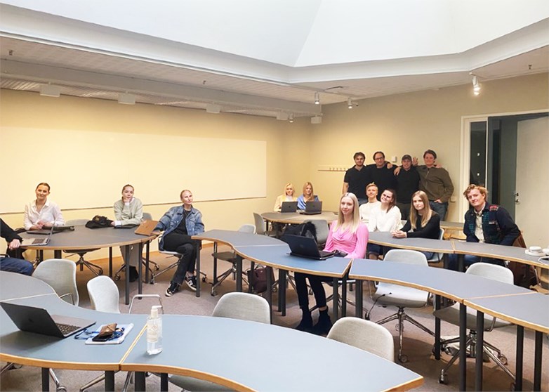 Students sit and stand spread out in a lecture room looking into the camera.