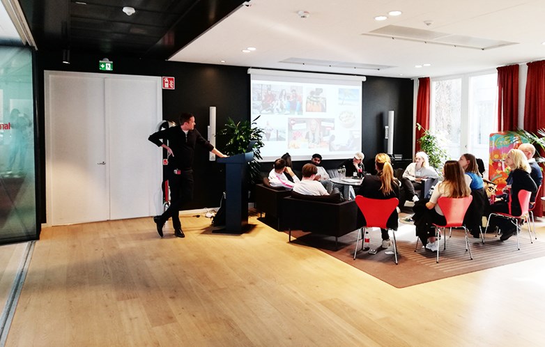 A representative from Coca-Cola hold a presentation for students sitting in cosy chairs in front a large projector screen.