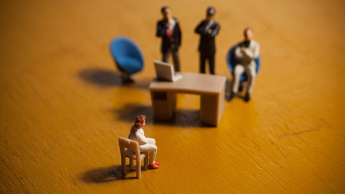Woman is interviewed by an intimidating group of business men. Concept of sexual harassment, intimidation, gender inequality or isolation. Depicted in miniature. Employee being fired or dismissed.