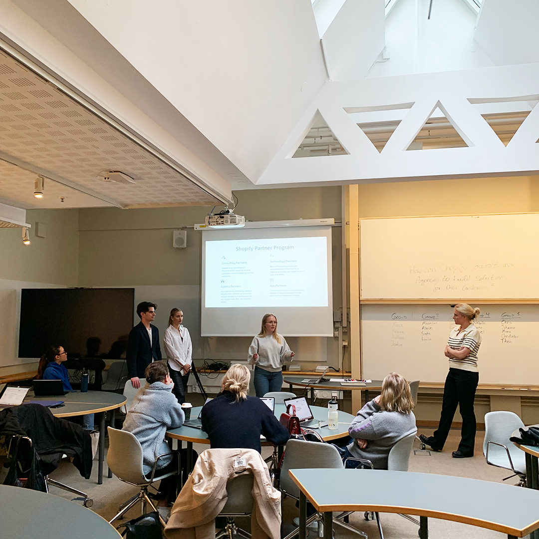 Students presenting in fron of a large TV screen in a high ceiling classroom.