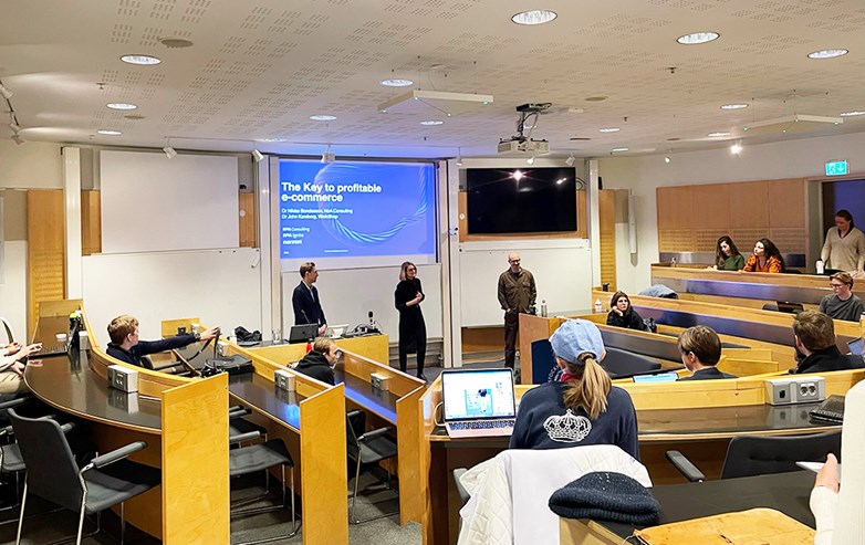 Three presenters standing in front of a class of students. "The Key to Profitable e-commerce" is showing on a powerpoint slide behind them.