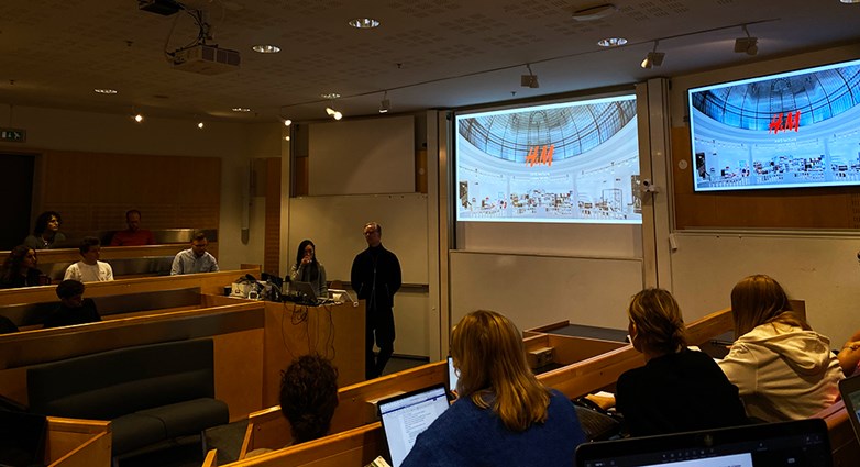 The reprentatives from H&M holds a presentation in front of students in a class room. The presentation is projected on two screens .