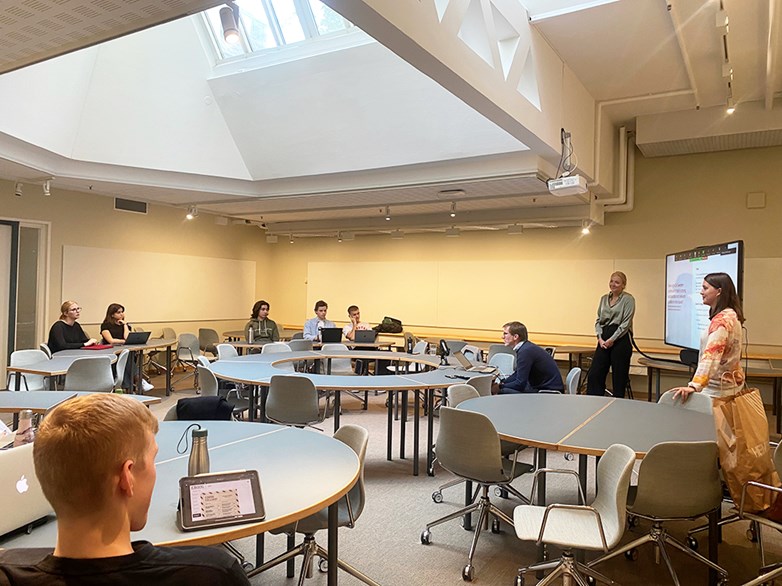 Students spread out in a lecture room sit and listen to two persons presenting in front a larger TV-screen.