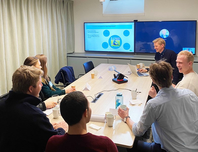 Seven persons sit around a table in a small conference room. One of them hold some kind of presentation projekted on two wall-fixed screens.