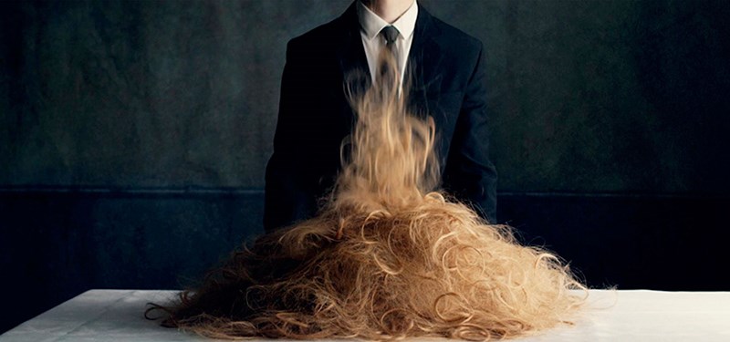A figure in a staged outdoor scene with long hair drawn across the face, concealing it entirely.