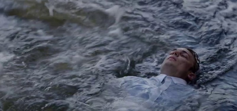 A young man floating on his back in dark water, face up and eyes open, clothed.