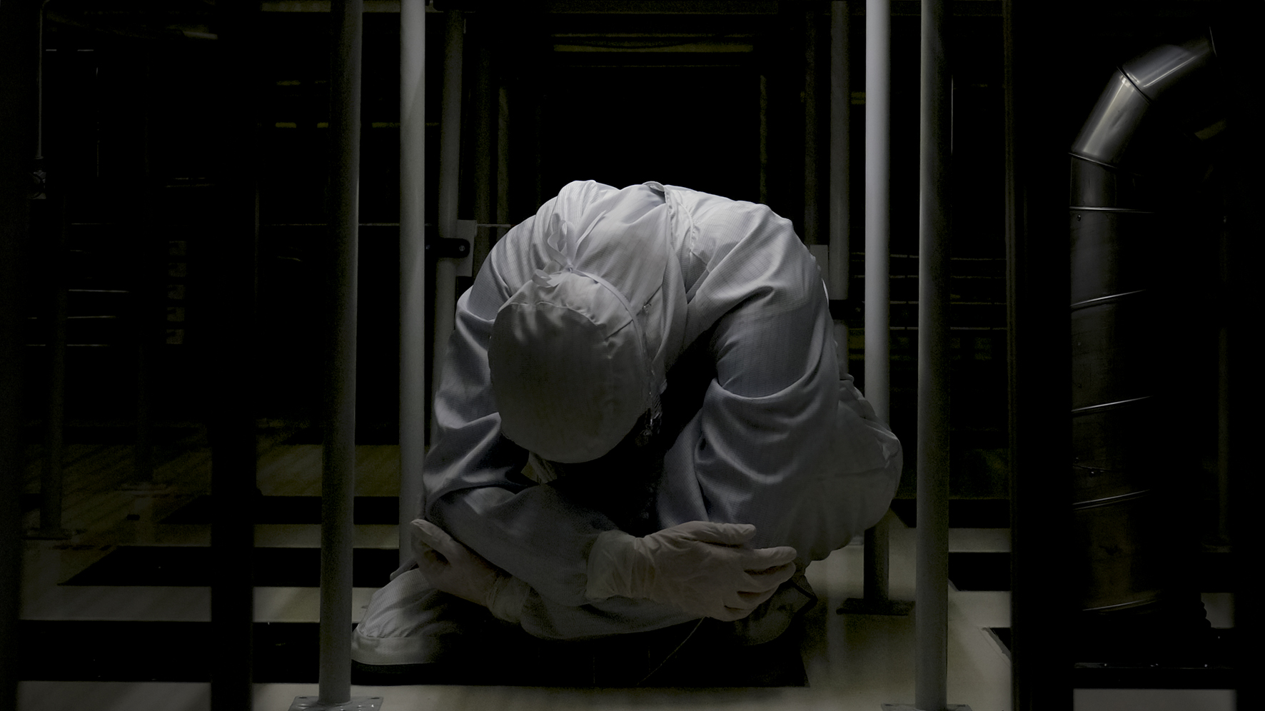 Researchers in full-body coveralls moving through a cleanroom under white and yellow light.