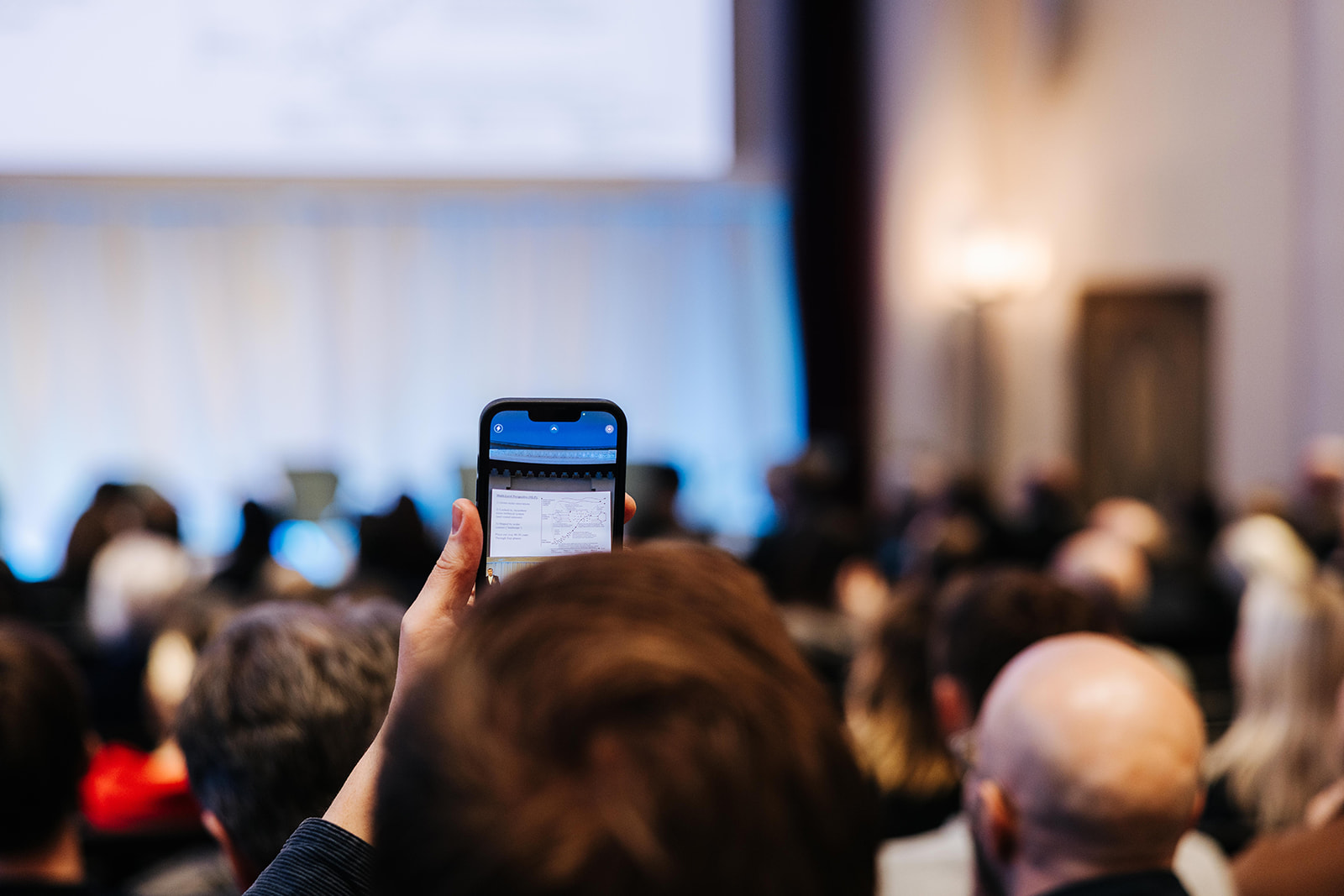 Woman sitting in a full auditorium, holding up her phone to take a picture and share updates from the event.