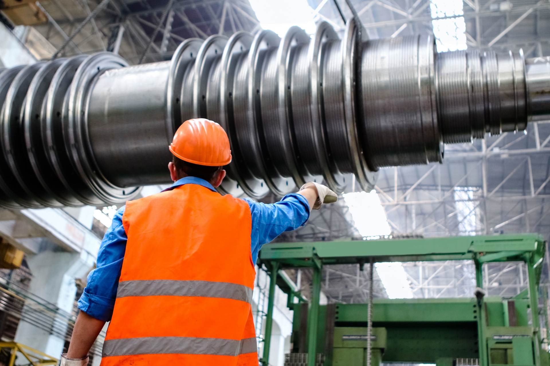 Worker in a steel factory, wearing an orange helmet and jacket, directing a colleague navigating large steel machinery.
