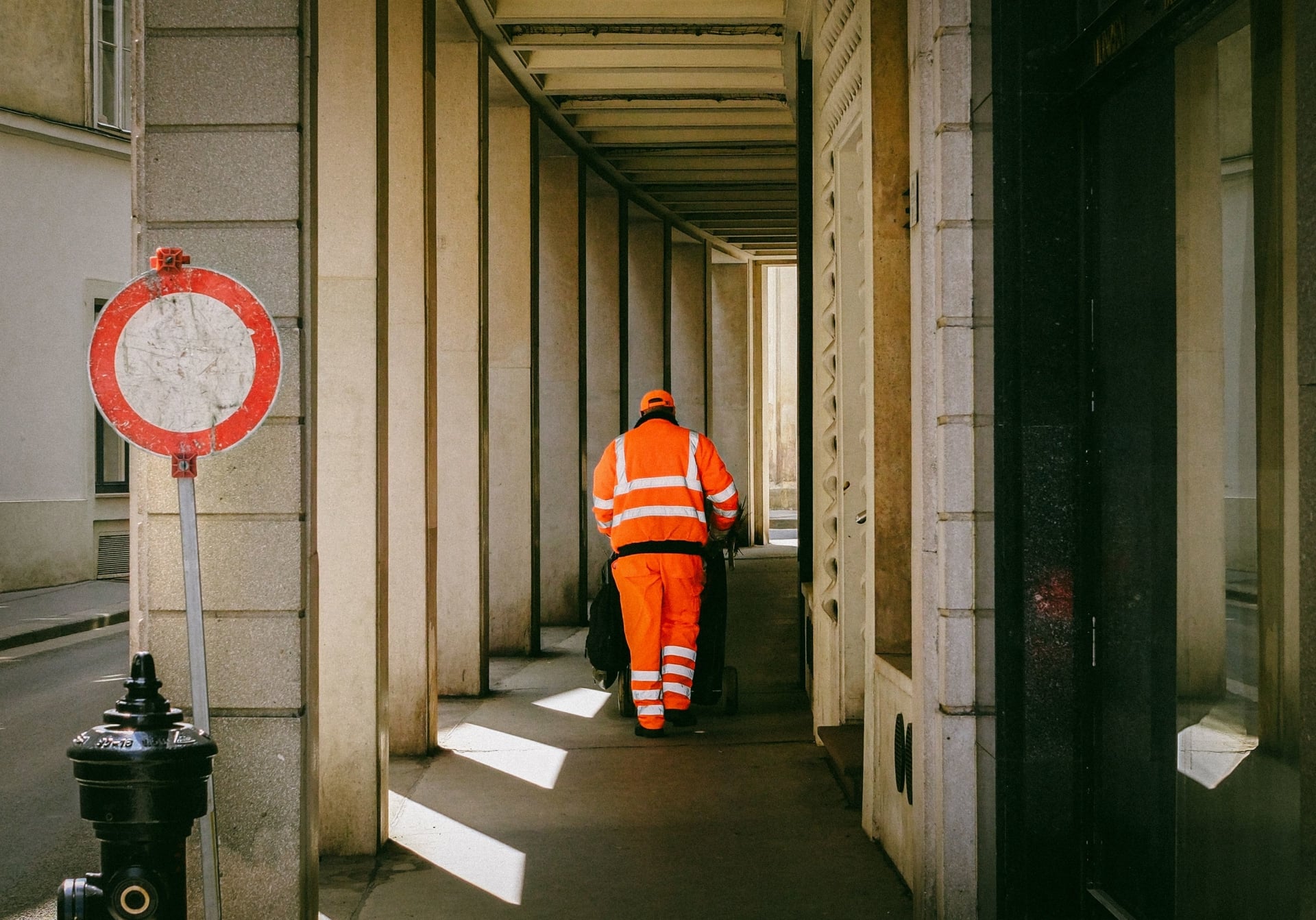 Public service worker walking down a street lined with columns, wearing an orange overall.