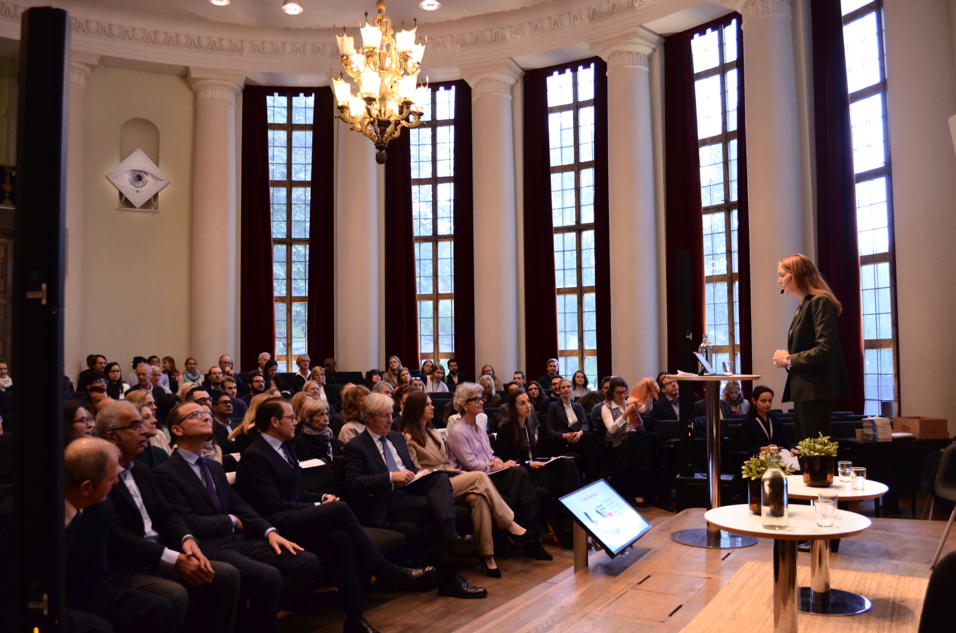 An auditorium full of people, listening to a speaker standing on stage. 