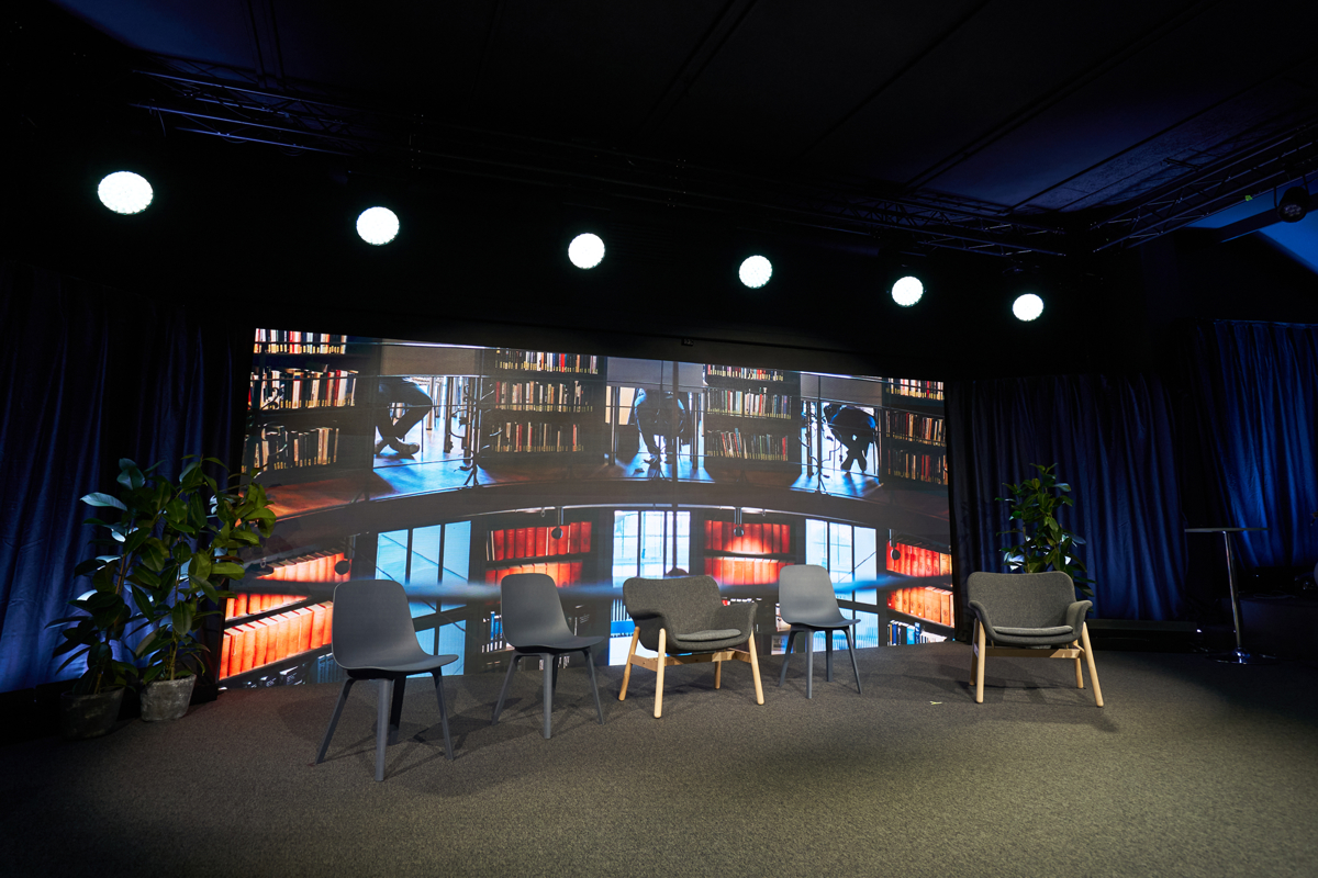 Five chairs lined up on a studio set, with a dark background with bookshelves.