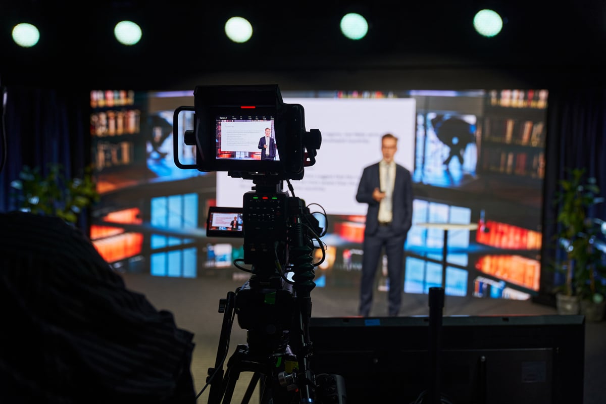 Camera filming a man standing on stage talking.