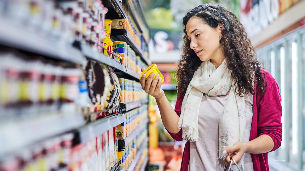 Woman shopping in a grocery store, holding up a yellow can and inspecting it.