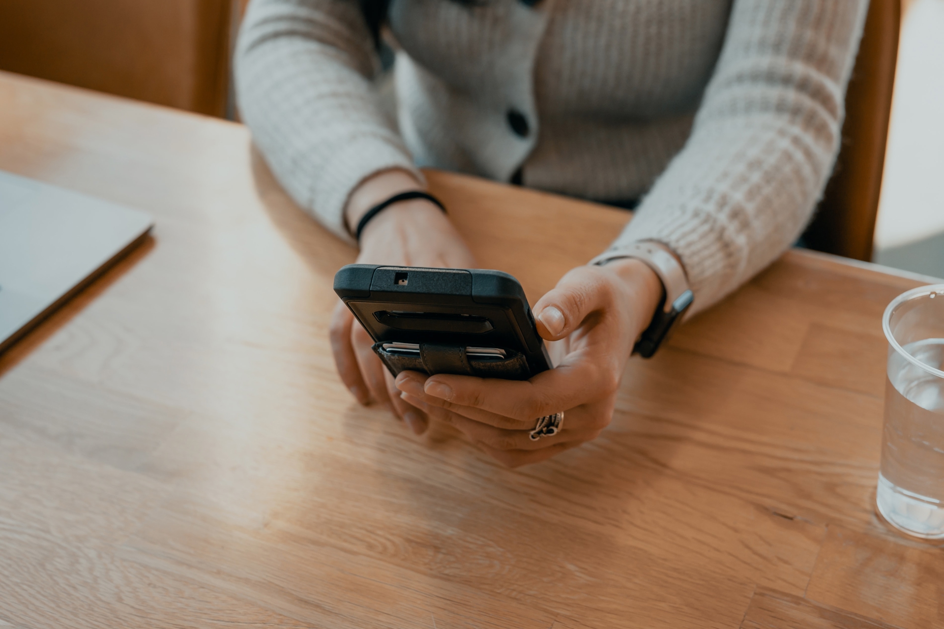 Woman in a grey sweater holding a phone in her hands with her arms on a desk. There's a glass of water to her left and a computer on her right.