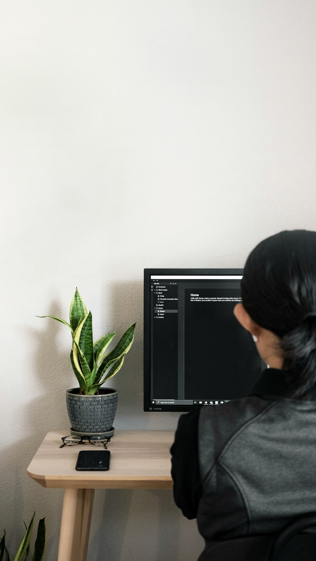 Woman sat in front of a computer, with her back to the camera. The walls are clean and white, and next to her on the desk is a plant.