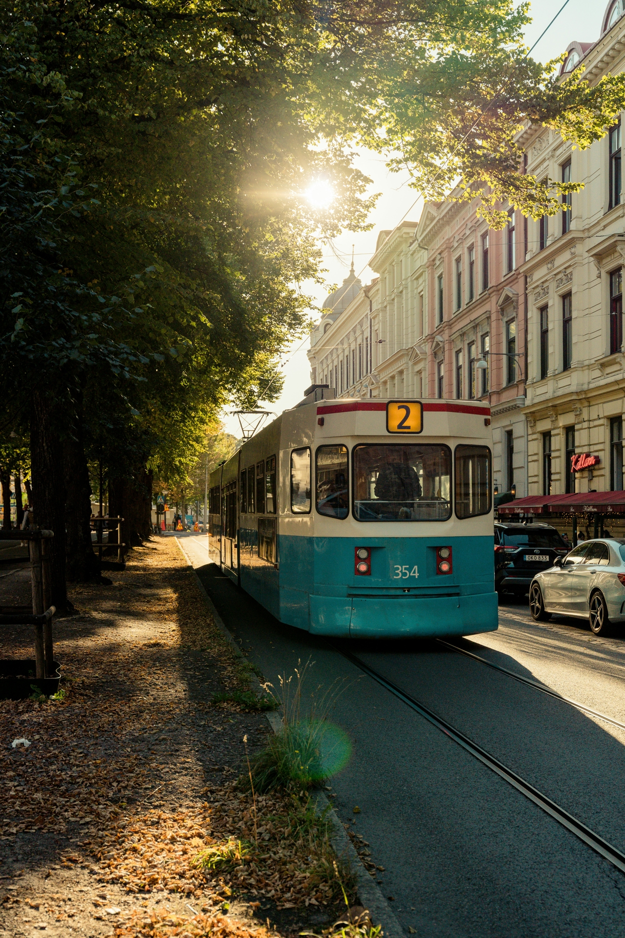 A tram in Sweden in an urban setting, passing by trees on one side and pavement and buildings on the other, with the sun in the background.