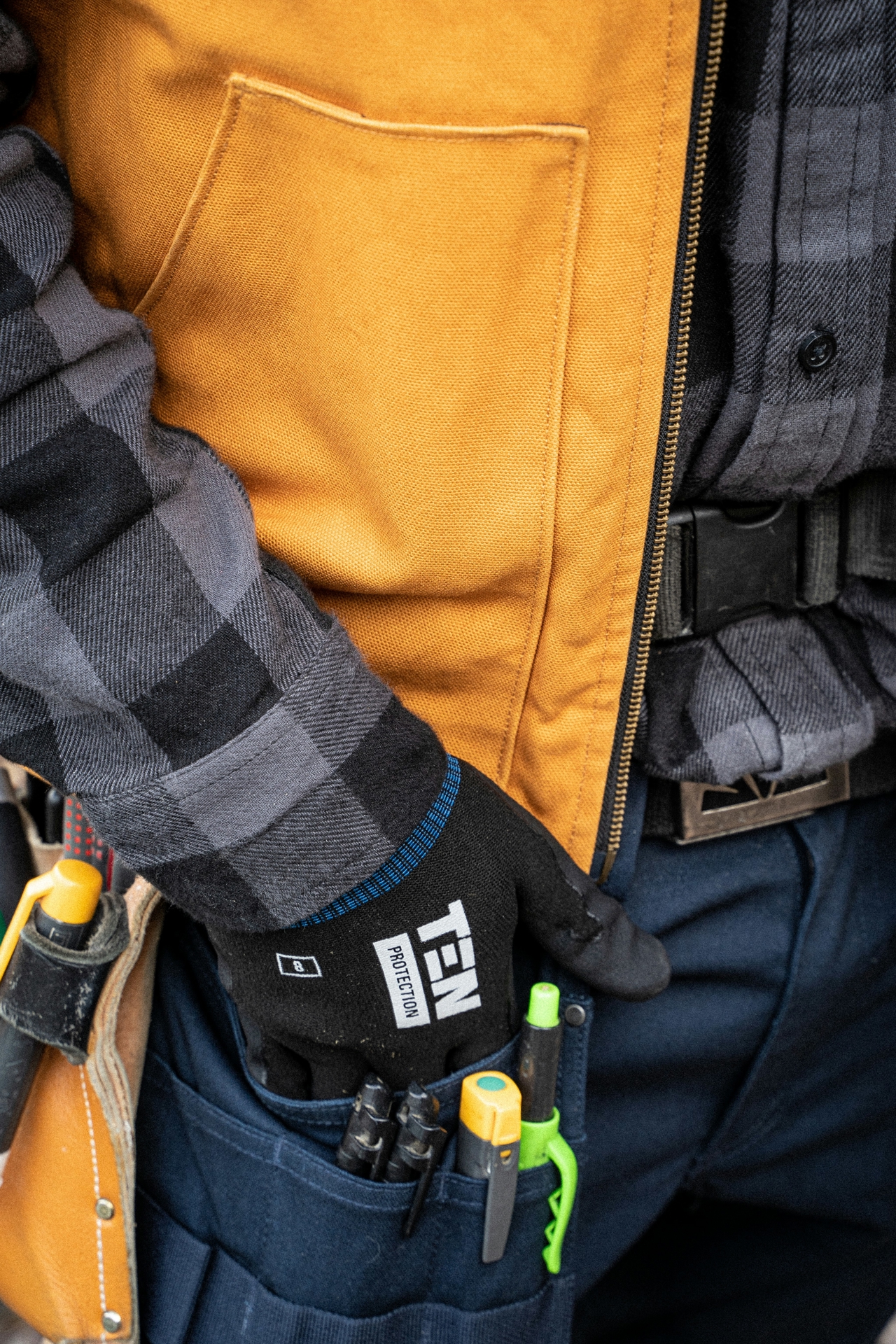 Close-up of a construction worker's outfit. Wearing a flannel shirt, yellow vest, black gloves and marine utility trousers.