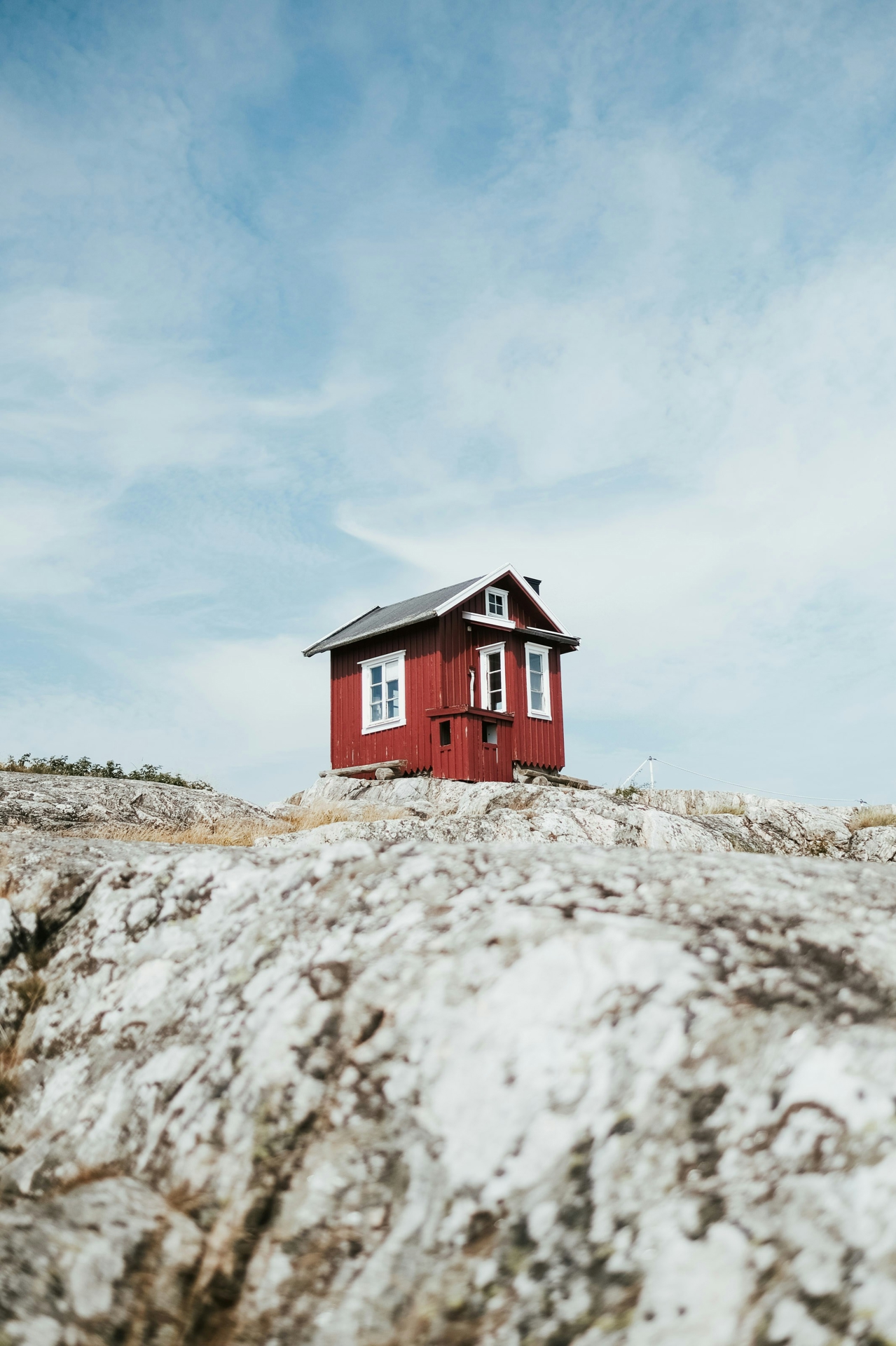A red Swedish cottage standing on coastal cliffs with a blue, somewhat cloudy, sky behind it.