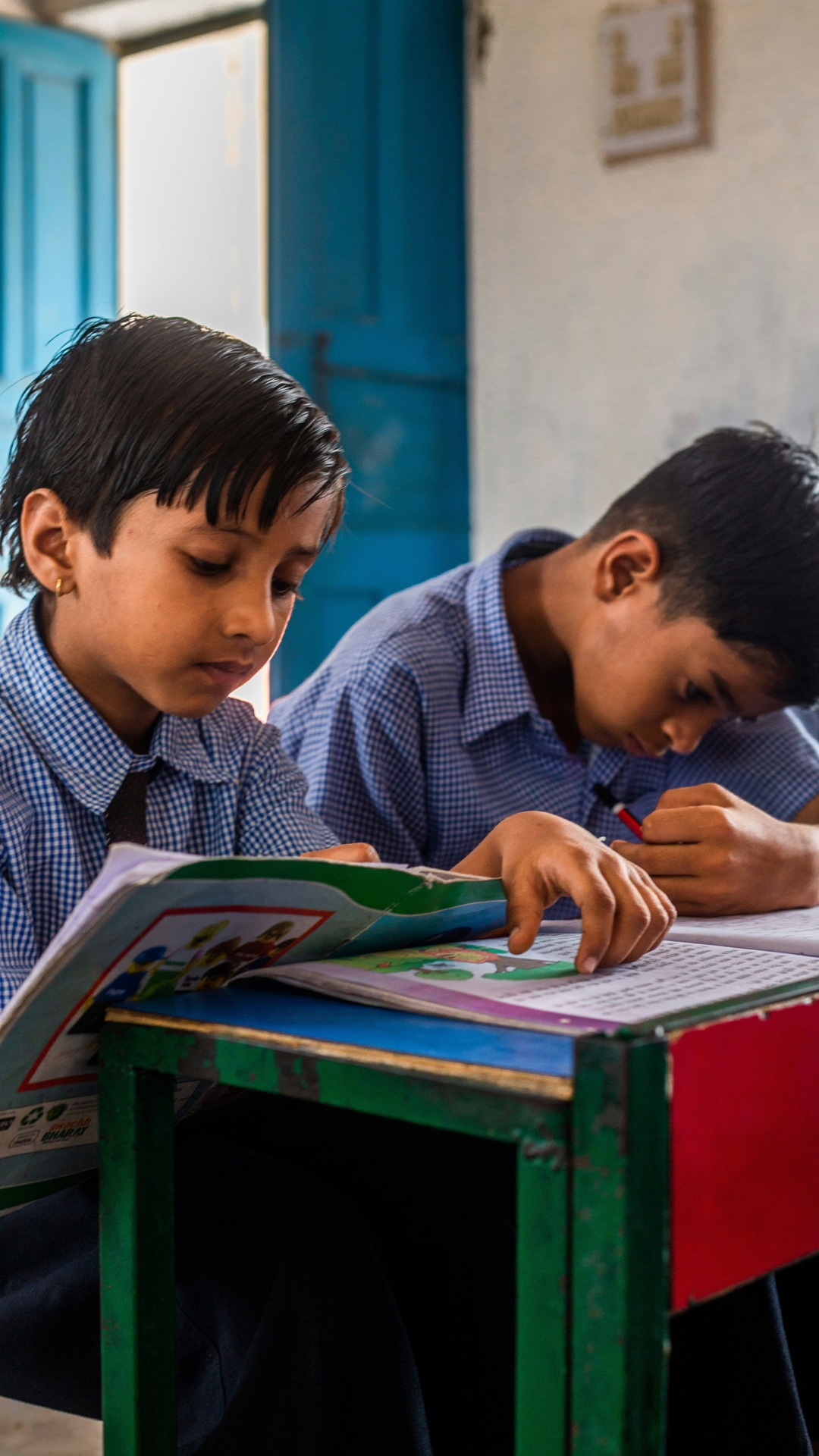 Two boys attending school in India, sitting by a green desk reading and writing.