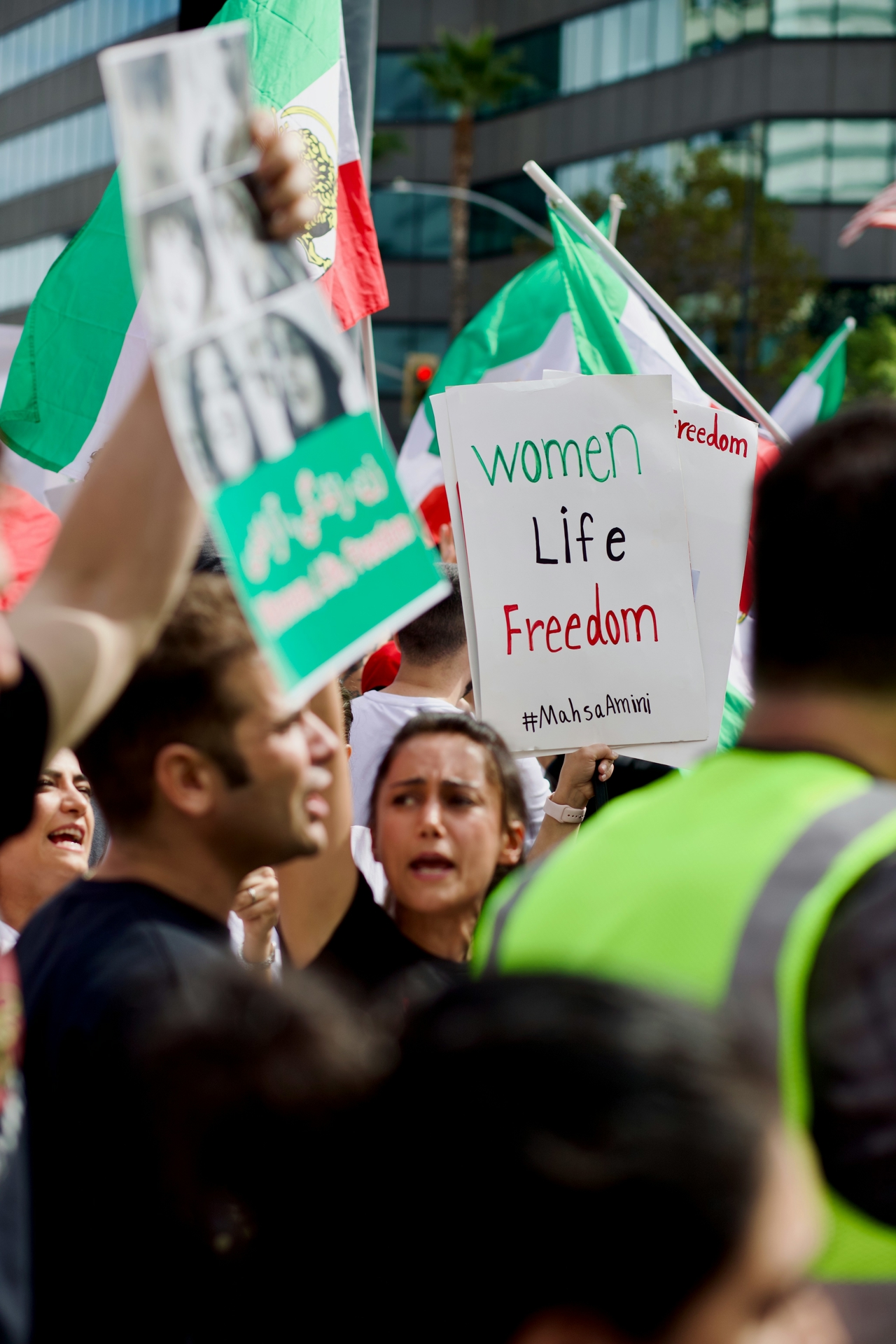 Woman standing in a crowd during a protest, holding a "Women, Life, Freedom" sign with the hashtag Mahsa Amini.