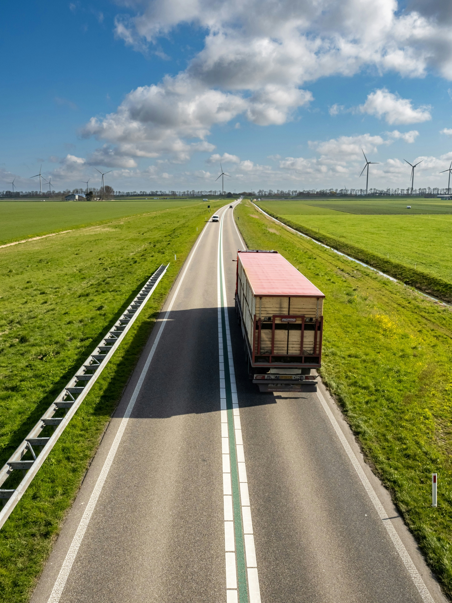 Freight truck driving on a road through green fields with wind turbines in the backdrop.