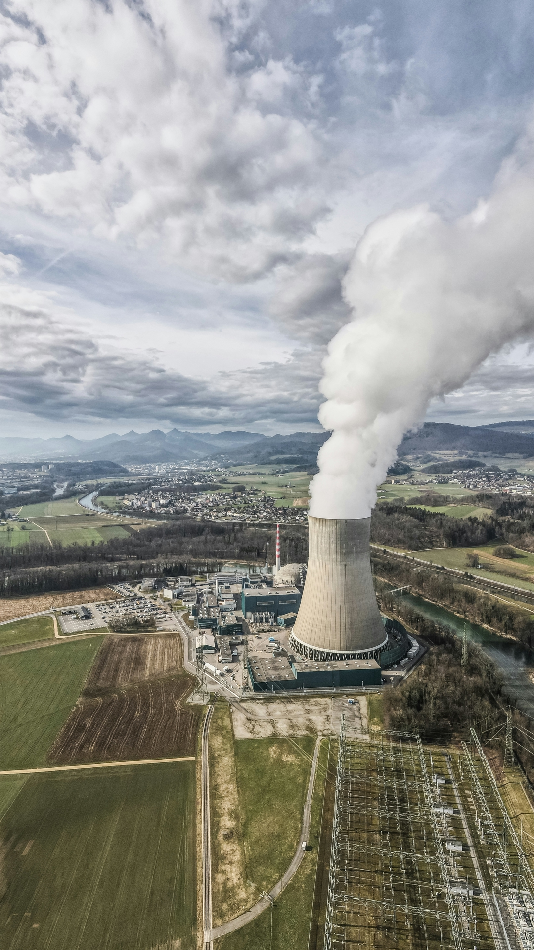 Industrial plant shot from the sky. Green fields surround the buildings spewing emissions, with mountains visible in the backdrop.